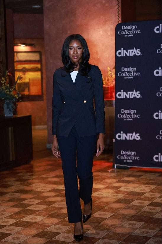 Female model in fitted navy blue pantsuit with gold buttons and white blouse, walking on indoor runway.