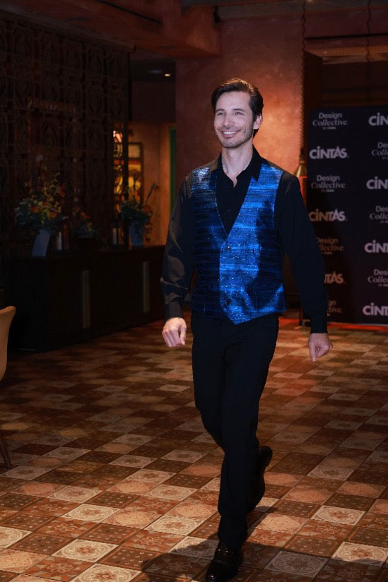 Male model smiling on the catwalk, wearing a black shirt and bright electric blue vest with black pants.